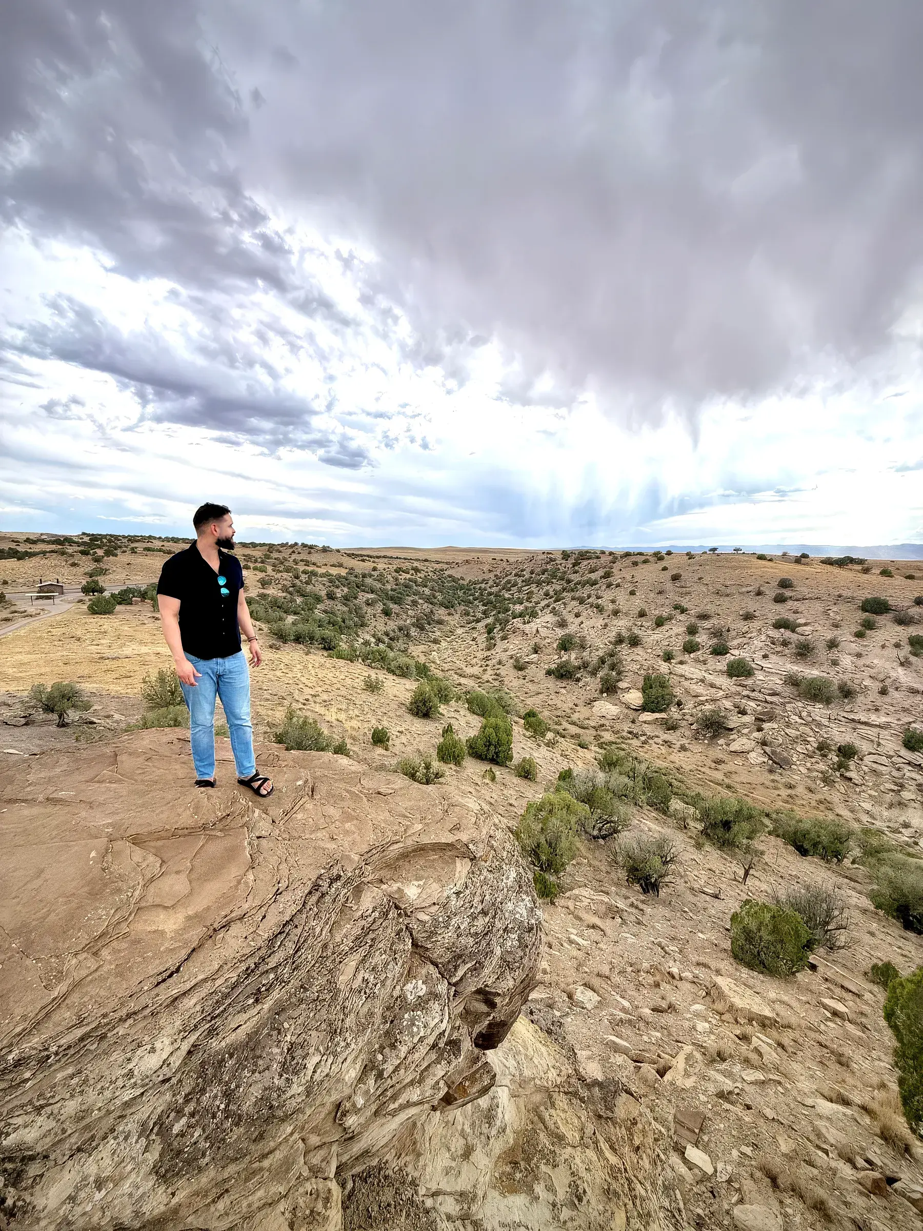 Johnson Gouvêa standing on a rock outcrop above a high-desert canyon, looking out towards the horizon under a heavy, clouded sky